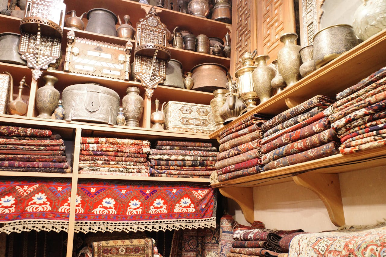 services-04 A vibrant scene showcasing folded Persian carpets and antique metalware in a traditional store setting.